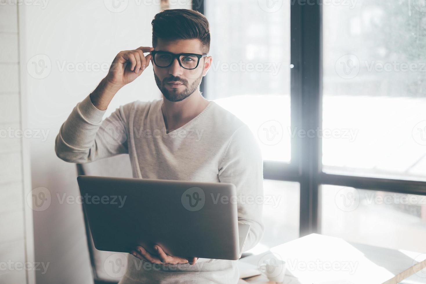 How may I help you  Confident young man holding laptop and adjusting his eyeglasses while leaning at the desk in office and looking at camera photo