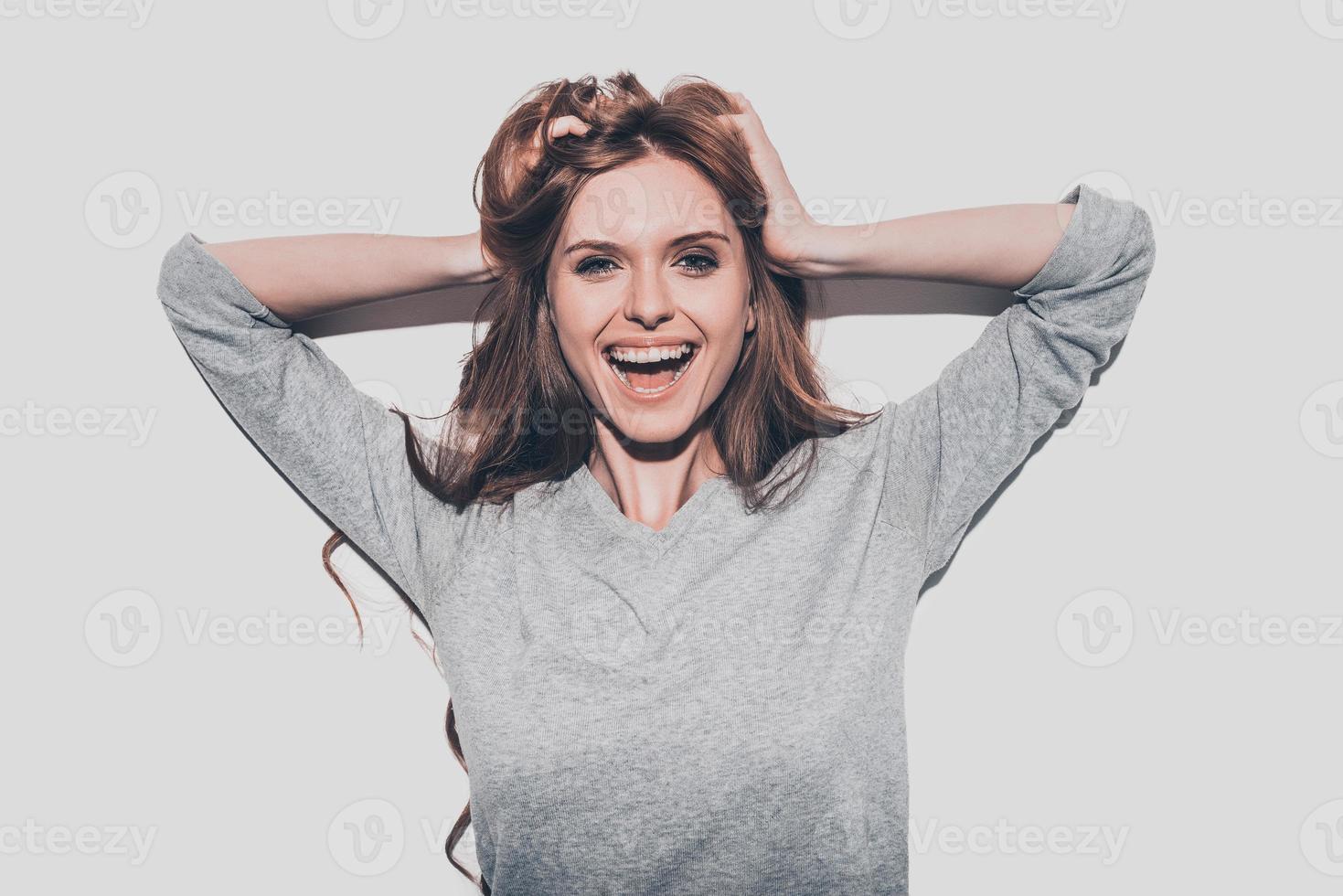 sintiéndose tan feliz, atractiva joven mujer sonriente cogida de la mano en el cabello y mirando a la cámara mientras está de pie contra un fondo gris foto