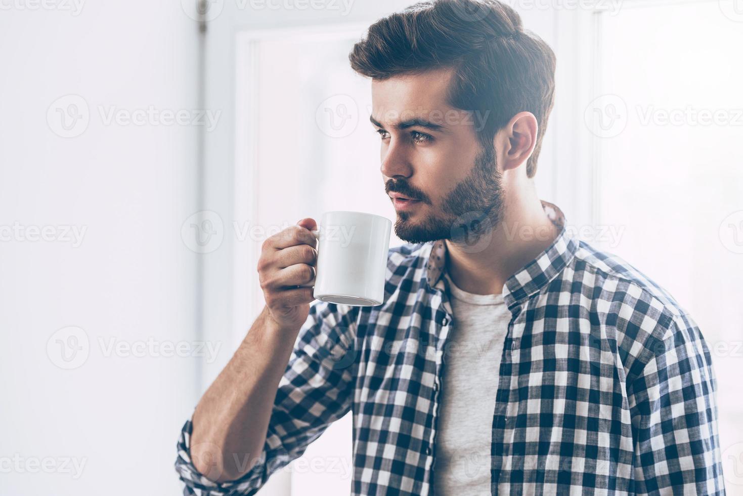 Inspired with cup of fresh coffee. Handsome young man holding coffee cup and looking thoughtful while standing in front of the window photo