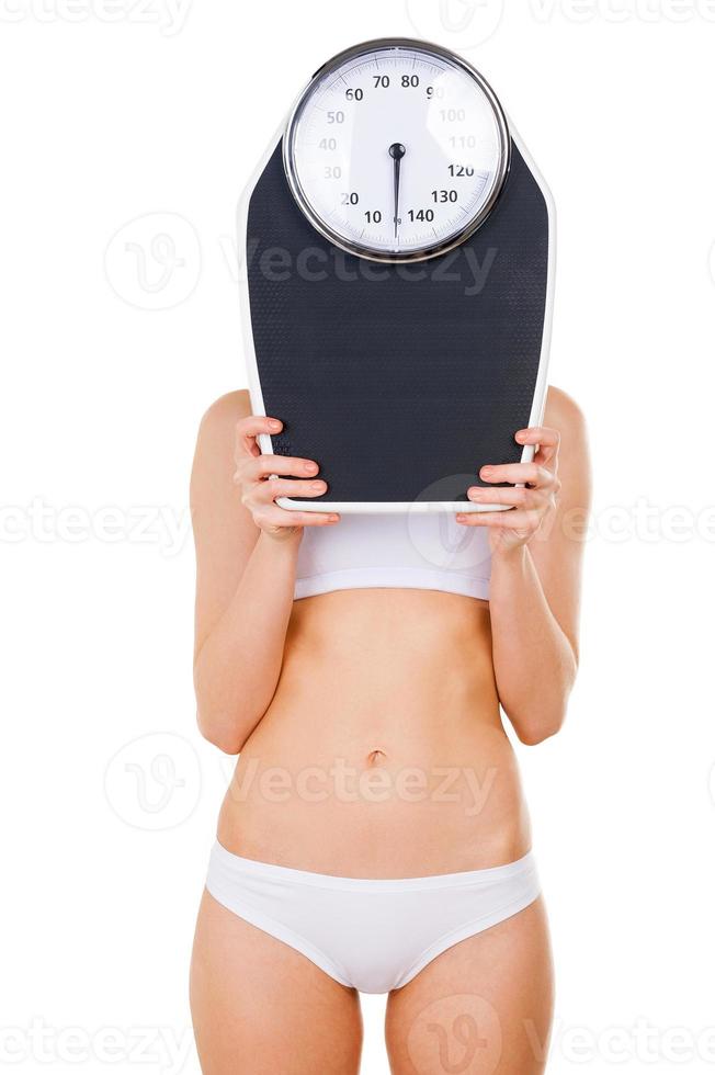 Woman with weight scale. Young slim woman in underwear hiding her face behind weight scale while standing isolated on white photo