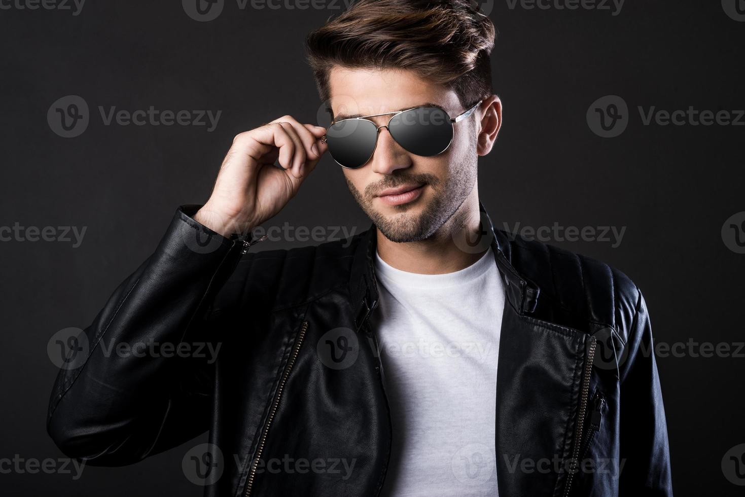 Cool and handsome. Handsome young man adjusting his sunglasses and looking at camera while standing against black background photo