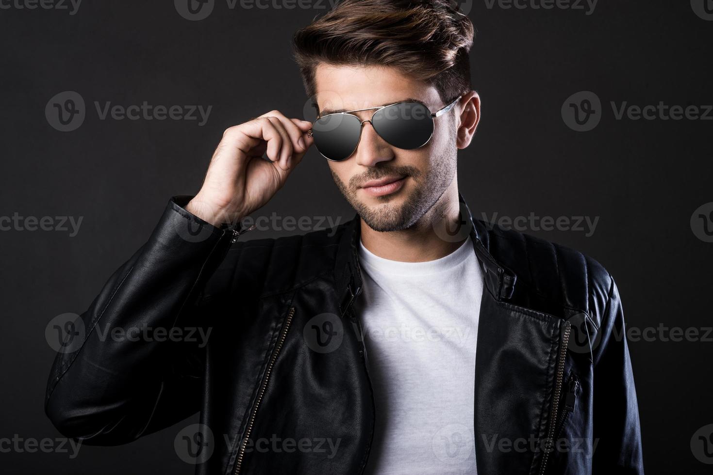 genial y guapo. apuesto joven ajustando sus gafas de sol y mirando a la cámara mientras está de pie contra el fondo negro foto