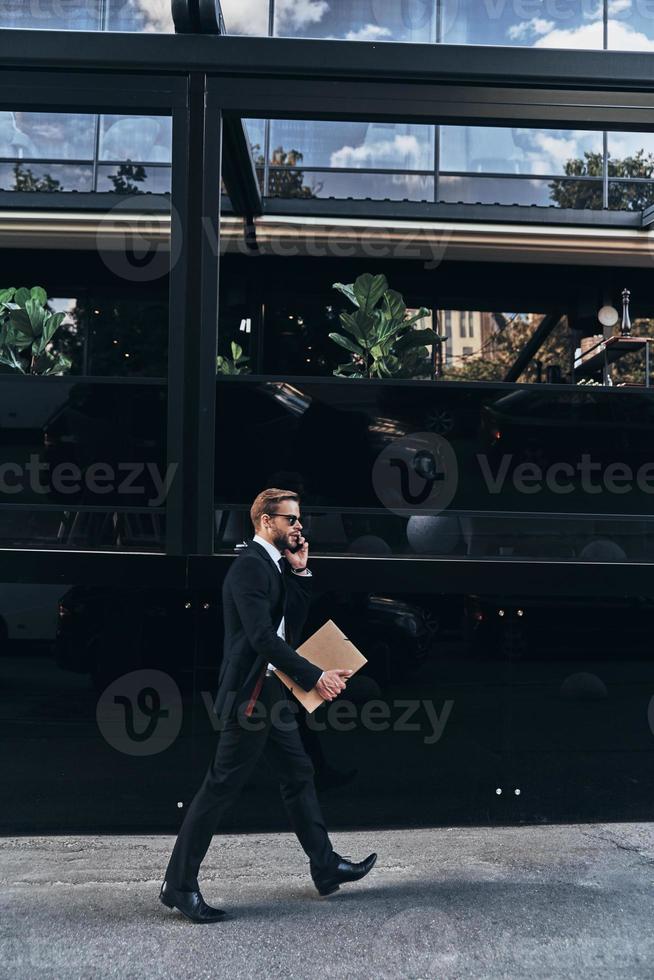 Always available. Full length of young man in full suit talking on the phone while walking outdoors photo