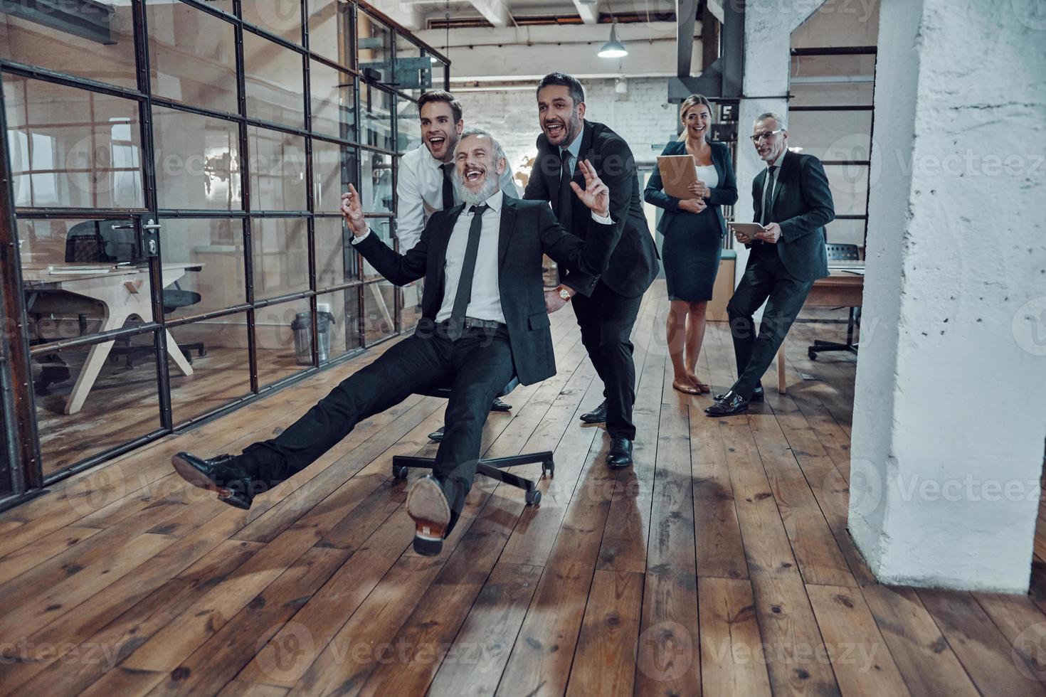 Playful businessmen pushing their boss on the office chair while running in the hallway photo