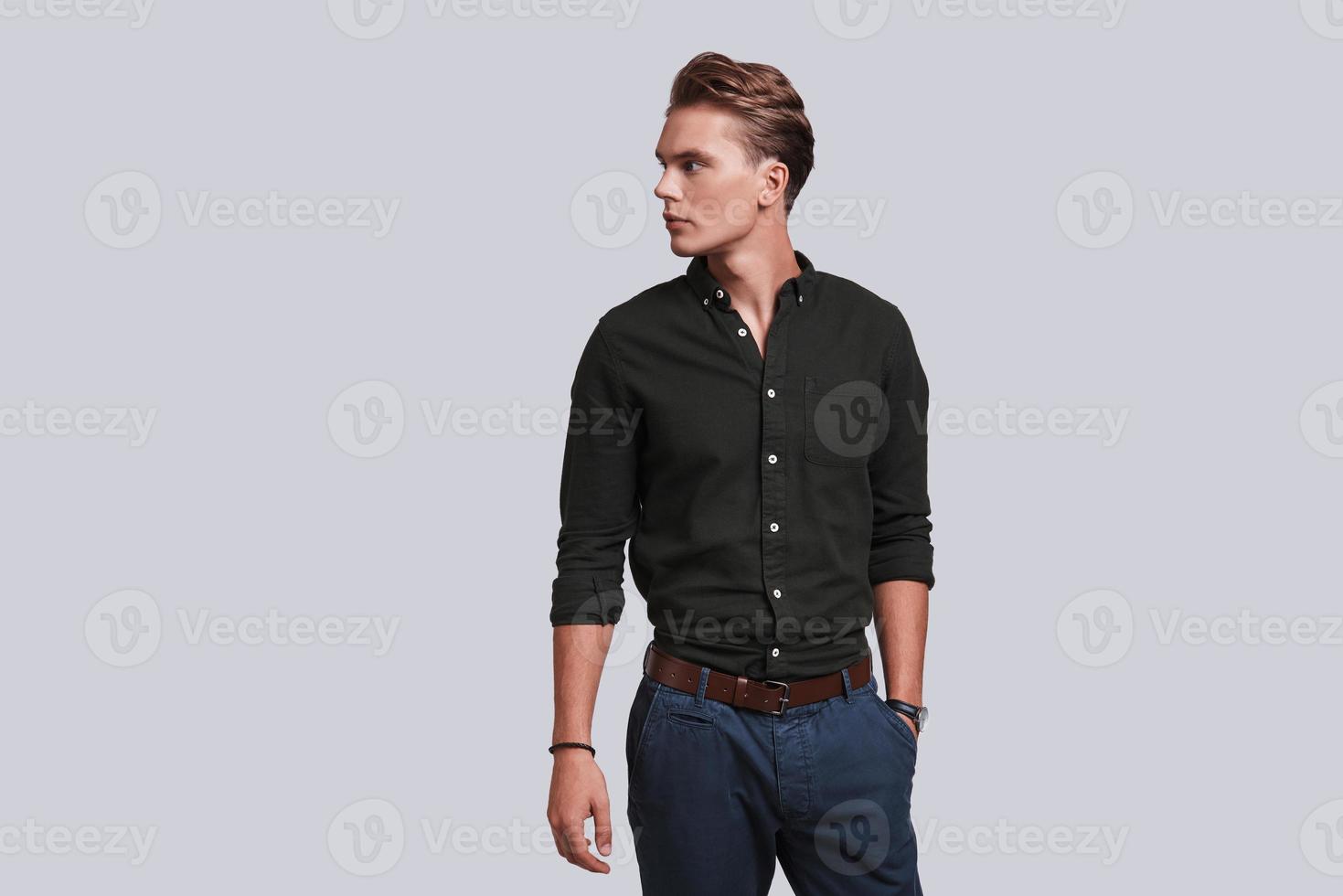 Confidence and charisma. Good looking young man keeping hand in pocket and looking away while standing against grey background photo