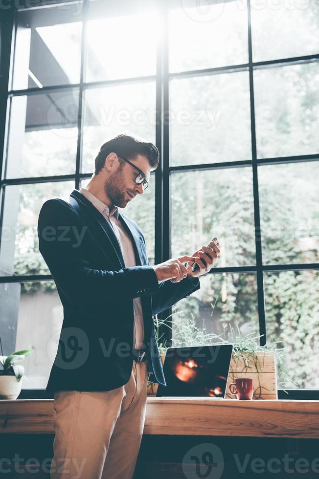Typing quick message to colleague. Low angle view of confident young man holding smart phone and looking at it while standing in front of the big window in office or cafe photo