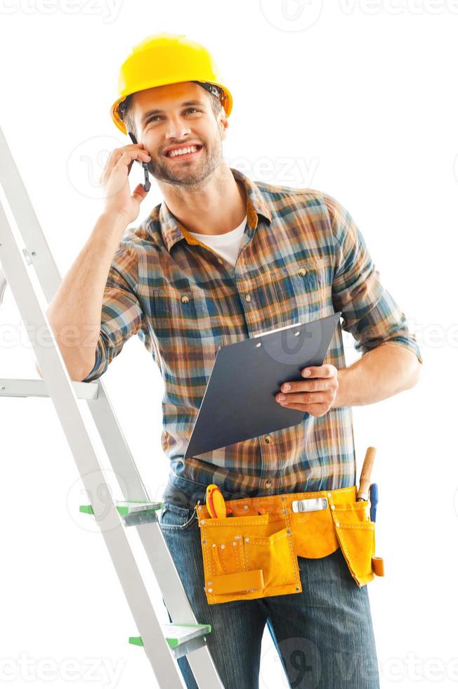Cheerful repairman. Smiling young manual worker talking on the mobile phone and holding clipboard while leaning at the ladder photo