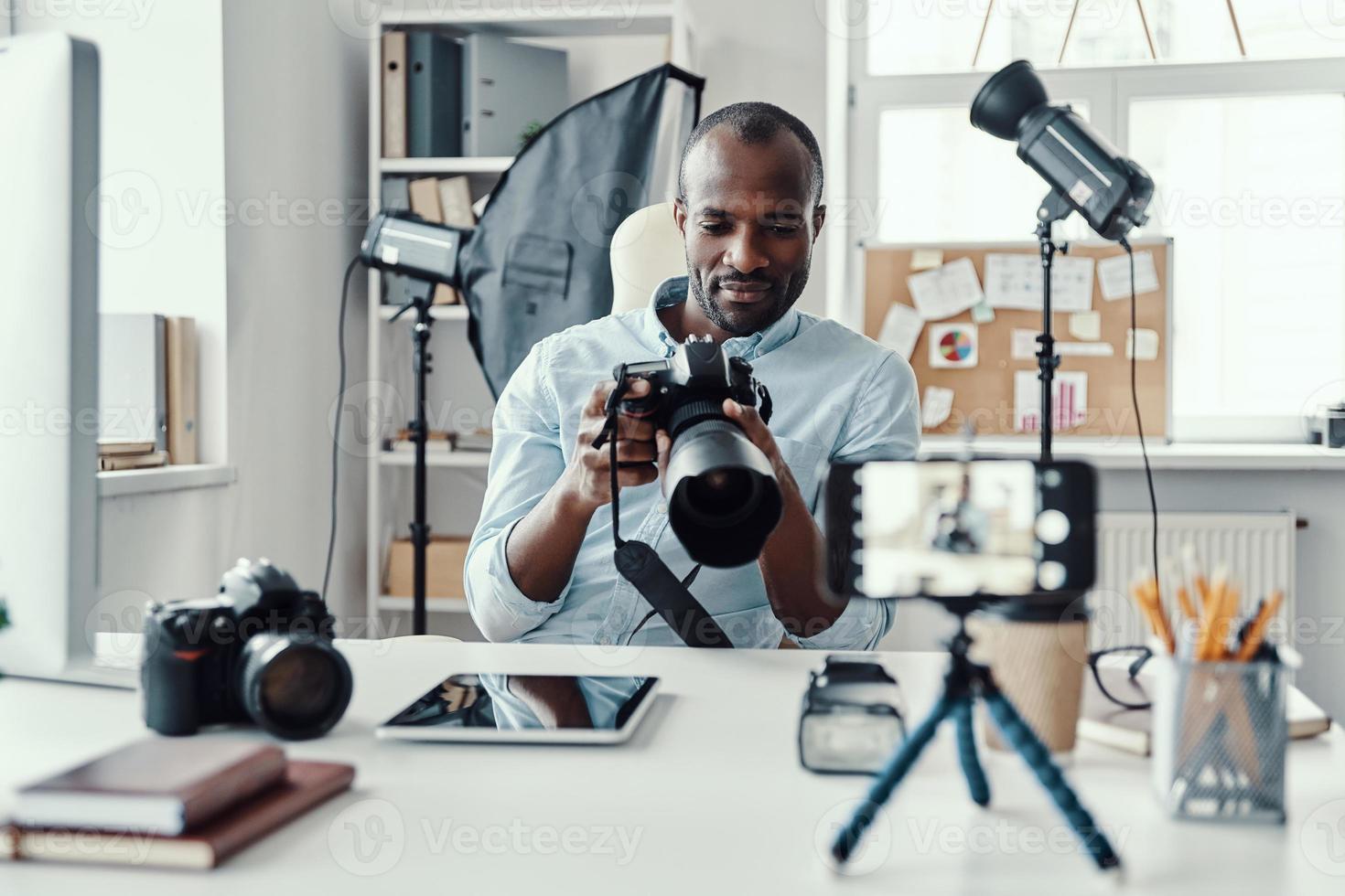 encantador joven africano con camisa que muestra una cámara digital y dice algo mientras hace un video en las redes sociales foto