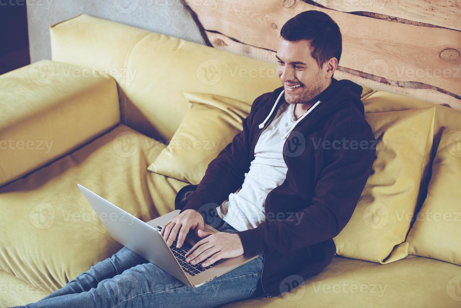 Surfing the net. Top view of cheerful young man using his laptop with smile while sitting on couch at home photo