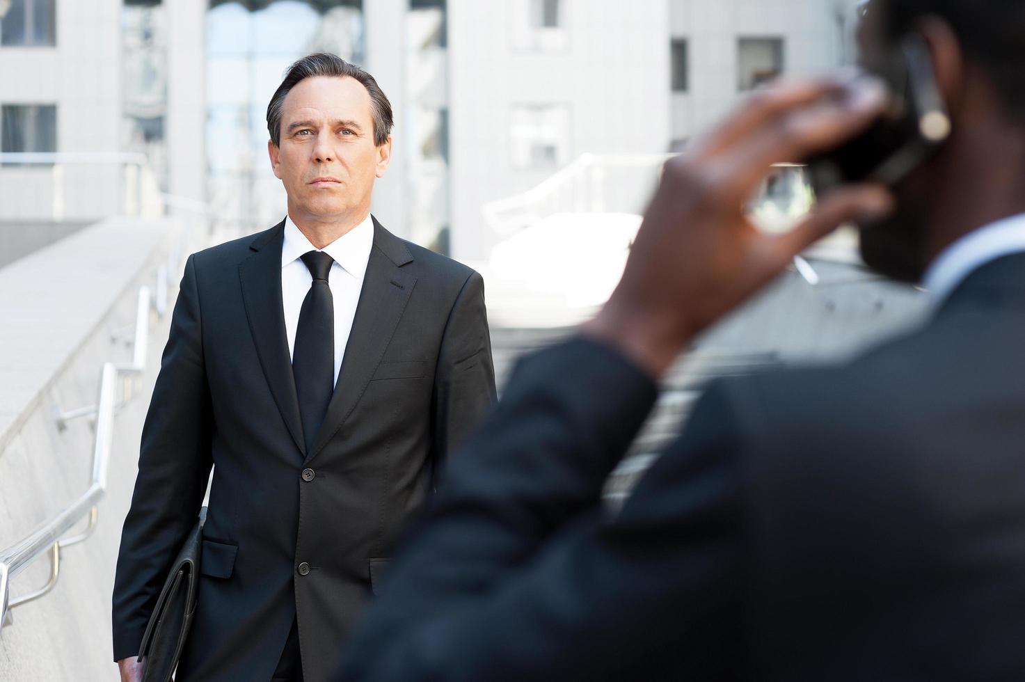 Confident business expert. Rear view of African man in formalwear talking on the mobile phone while another businessman walking by stairs on background photo