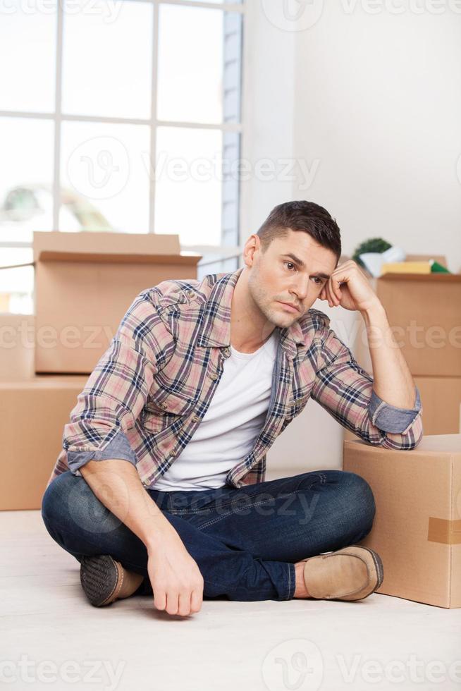 So much work ahead. Depressed young man sitting on the floor and leaning at the carton box while more cardboard boxes laying on the background photo