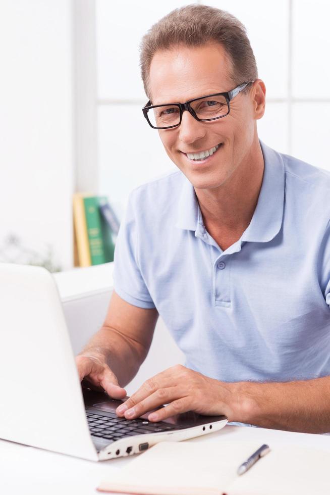 Enjoying working at home. Confident mature man working on laptop and smiling while sitting on the couch at home photo