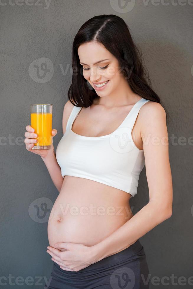 Fresh juice for my baby. Beautiful pregnant woman holding a glass of orange juice and smiling while standing against grey background photo