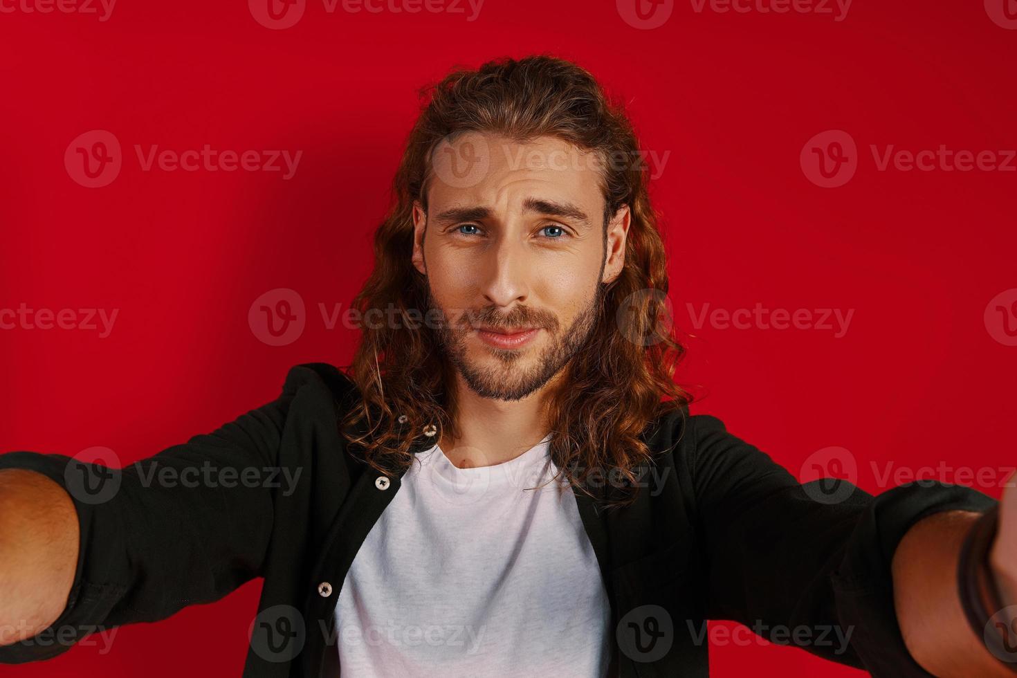 Self portrait of handsome young man in casual clothing looking at camera and smiling while standing against red background photo