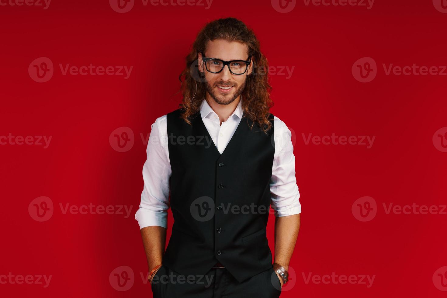 Charming young man in elegant clothing looking at camera and smiling while standing against red background photo