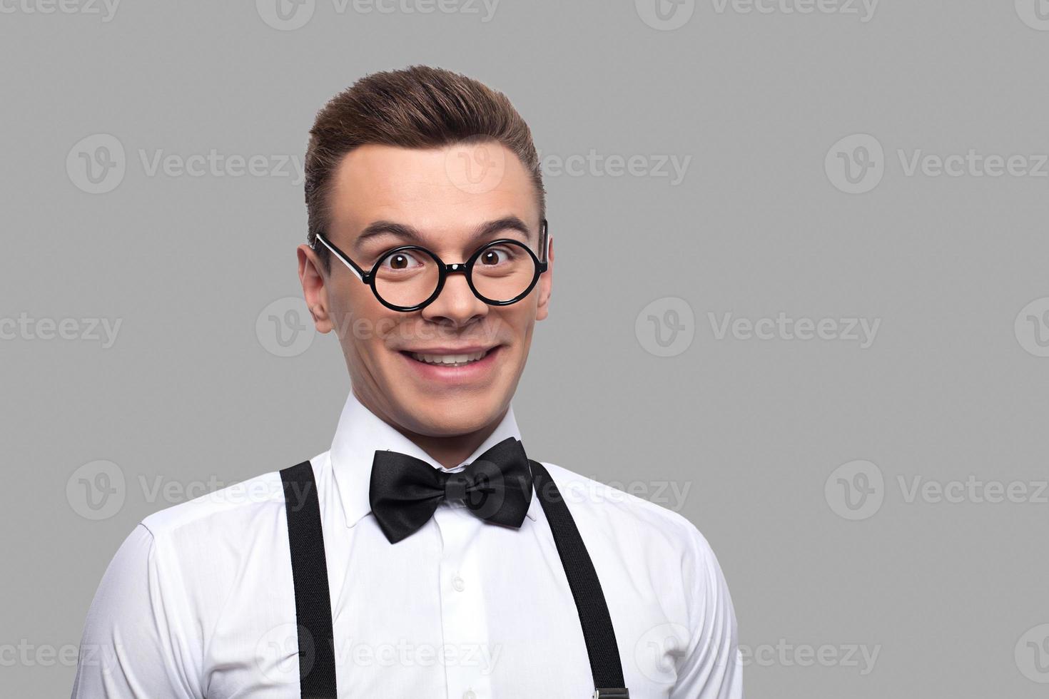 I know a lot of things. Portrait of young nerd man in bow tie and suspenders looking at camera and smiling while standing against grey background photo