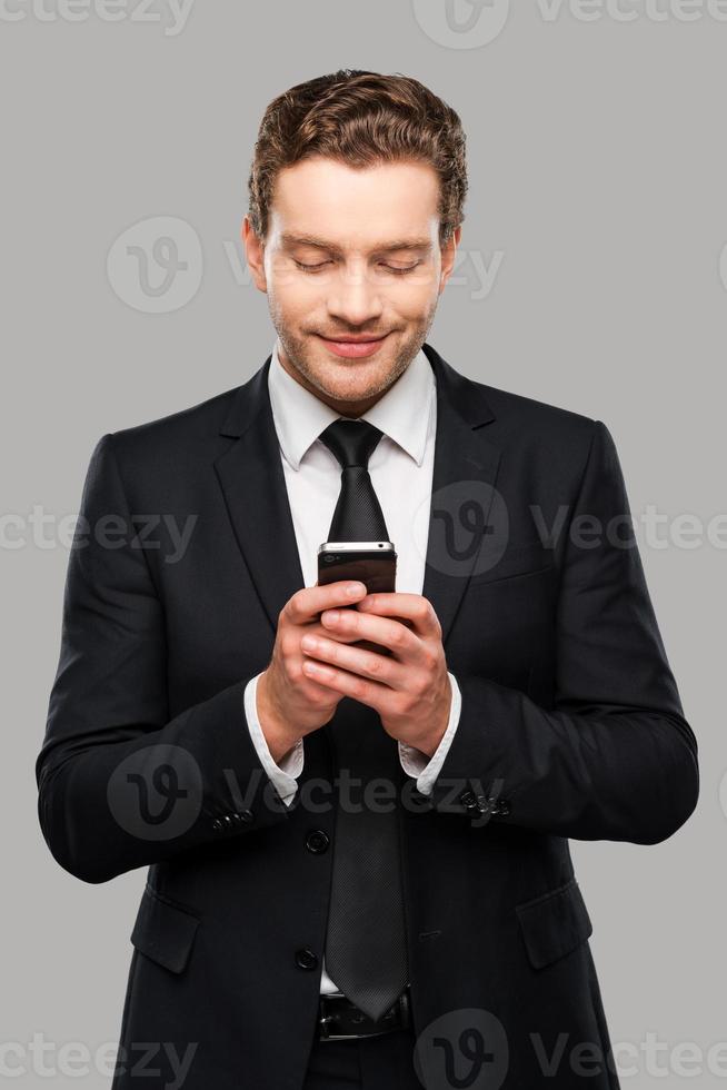 Typing business message. Confident young man in formalwear holding mobile phone and smiling while standing against grey background photo