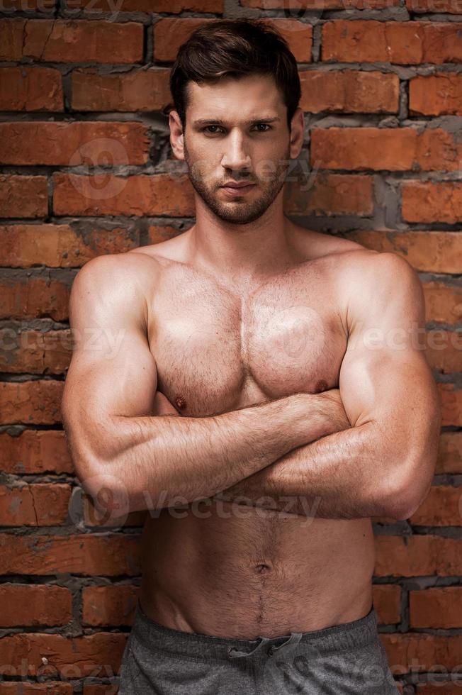 Strong man. Confident young muscular man keeping arms crossed and looking at camera while standing against brick wall photo