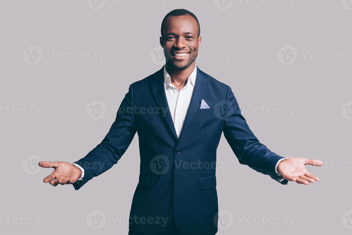 You are welcome Handsome young African man in smart casual jacket gesturing and smiling while standing against grey background photo