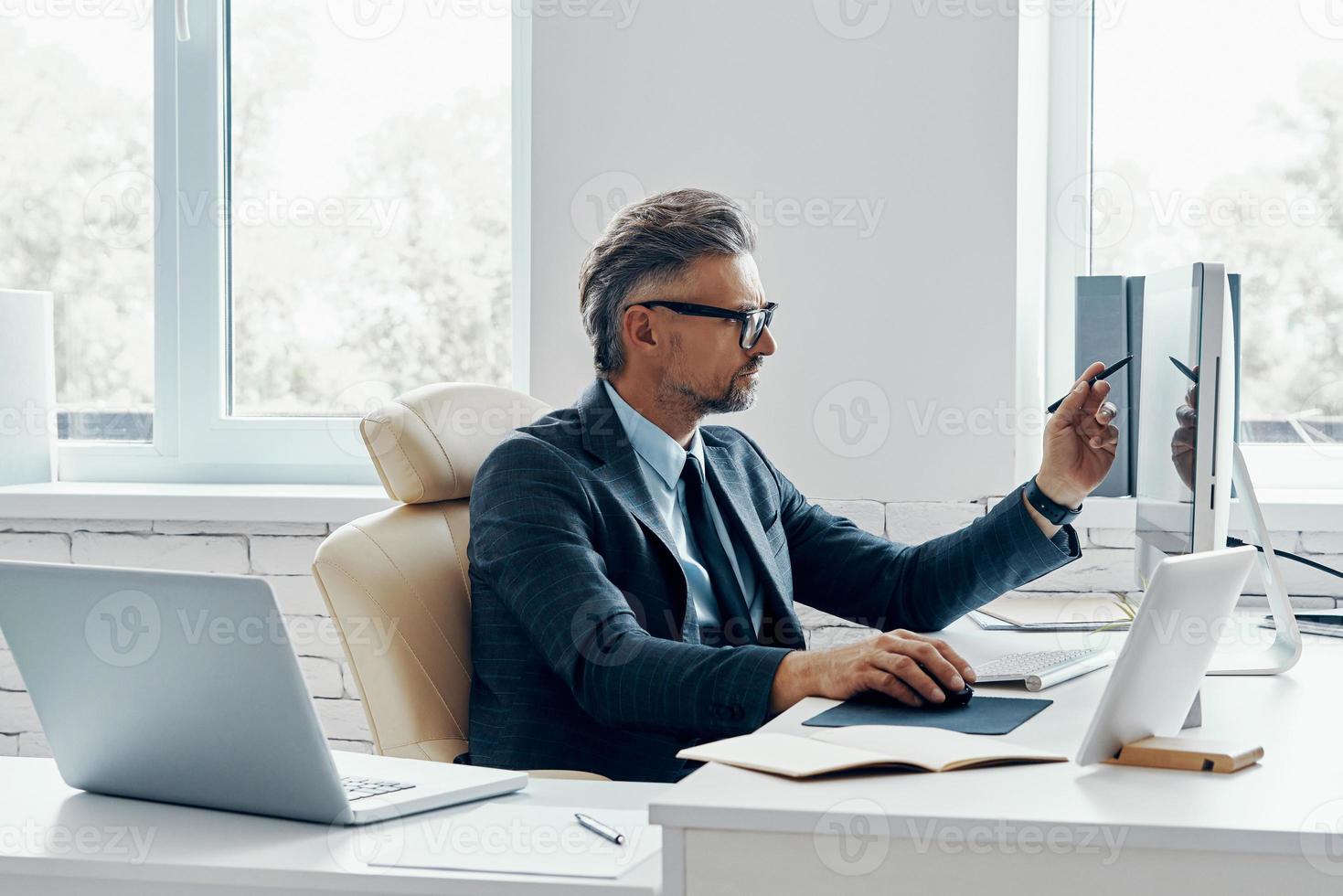 Confident mature businessman pointing computer monitor while sitting at his working place photo