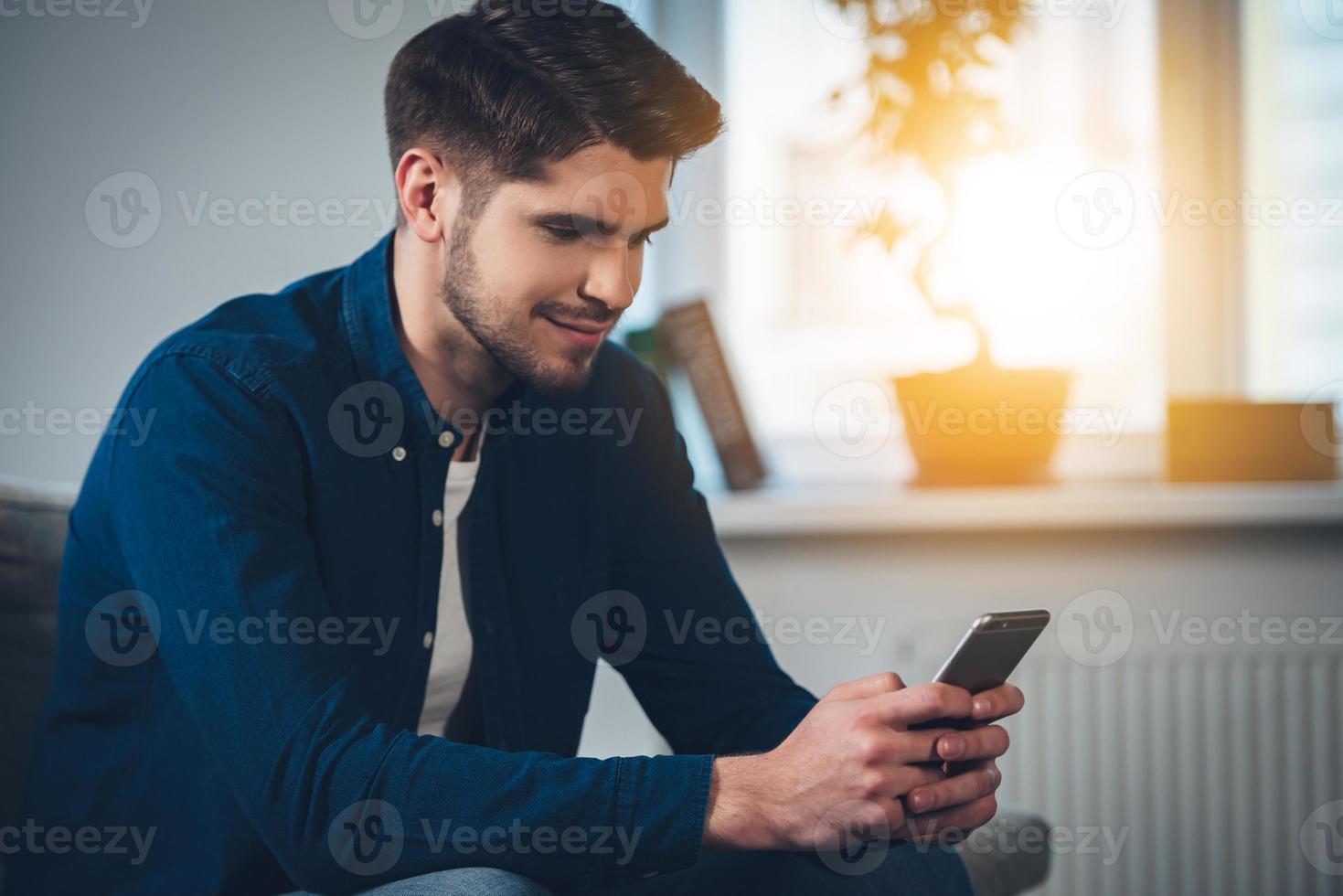 Lovely message from her. Side view of handsome young man using his smartphone with smile while sitting on the couch at home photo