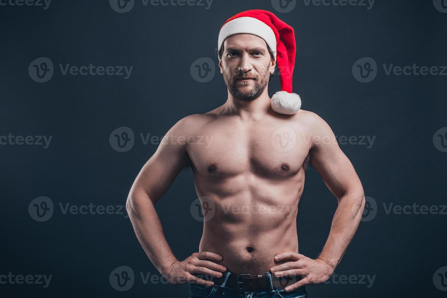 Thinking about Christmas presents. Young confident man keeping hands on hip and looking at camera while standing against black background photo