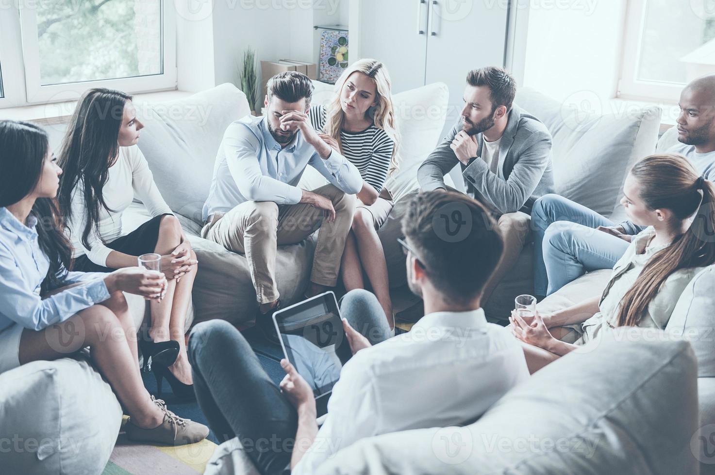 Everything will be alright. Group of young people sitting in circle while one man holding head in hand and looking upset while young woman comforting him photo
