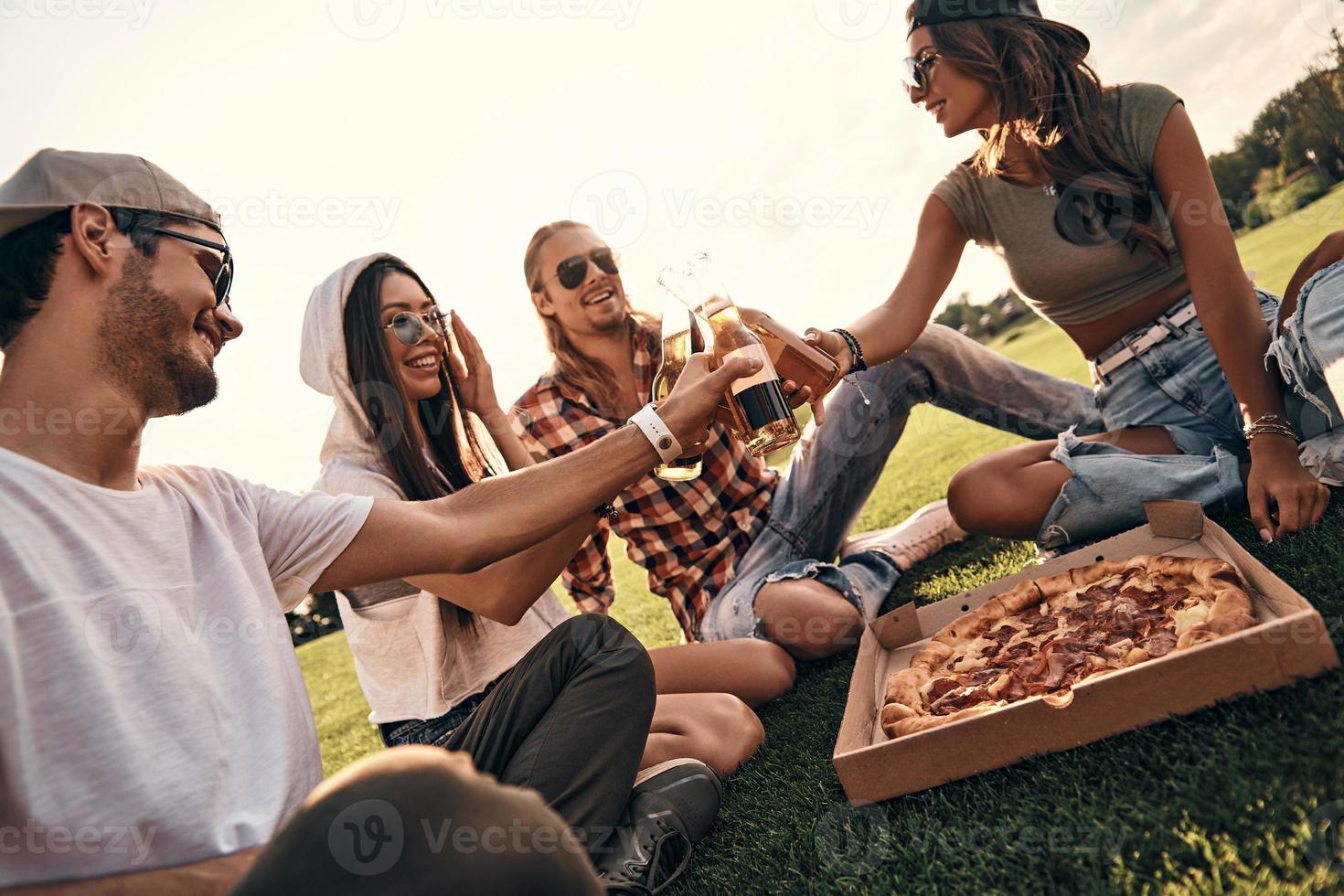 joven y despreocupado. grupo de personas sonrientes con ropa informal brindando con botellas de cerveza mientras disfrutan de un picnic al aire libre foto