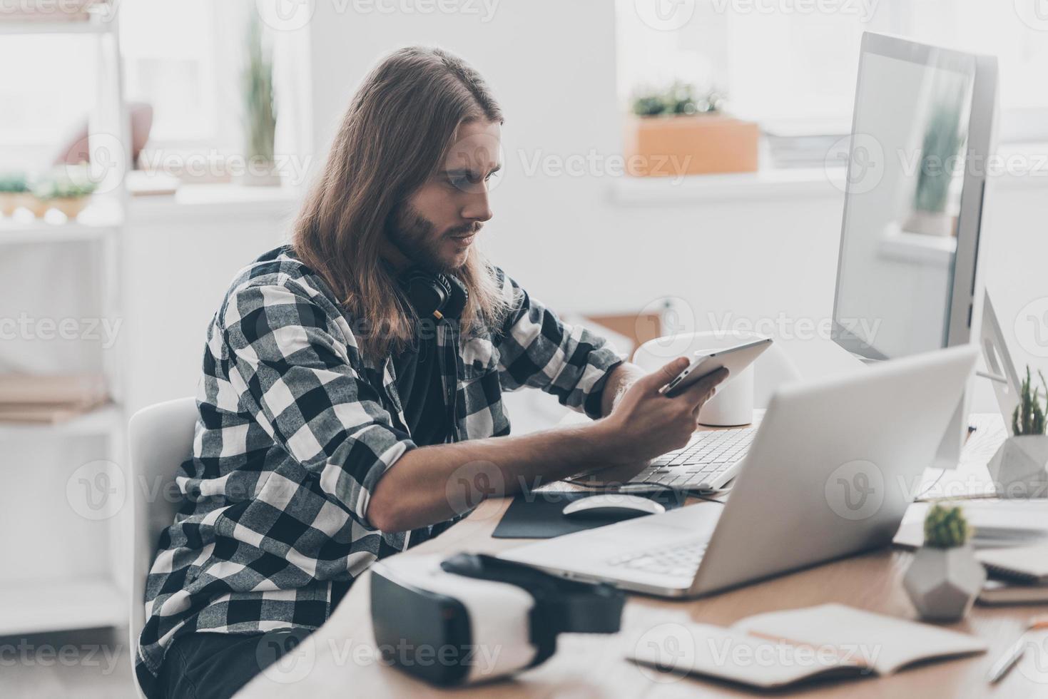 Busy working day. Handsome young man with long hair working on the digital tablet while sitting at his desk in creative office photo