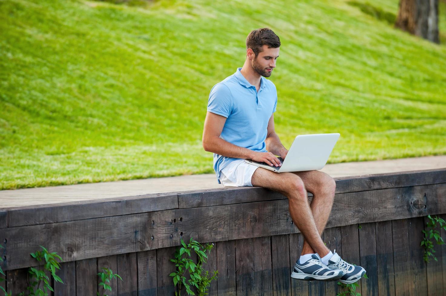 Surfing web wherever he wants. Handsome young man in polo shirt working on laptop while sitting on quayside photo