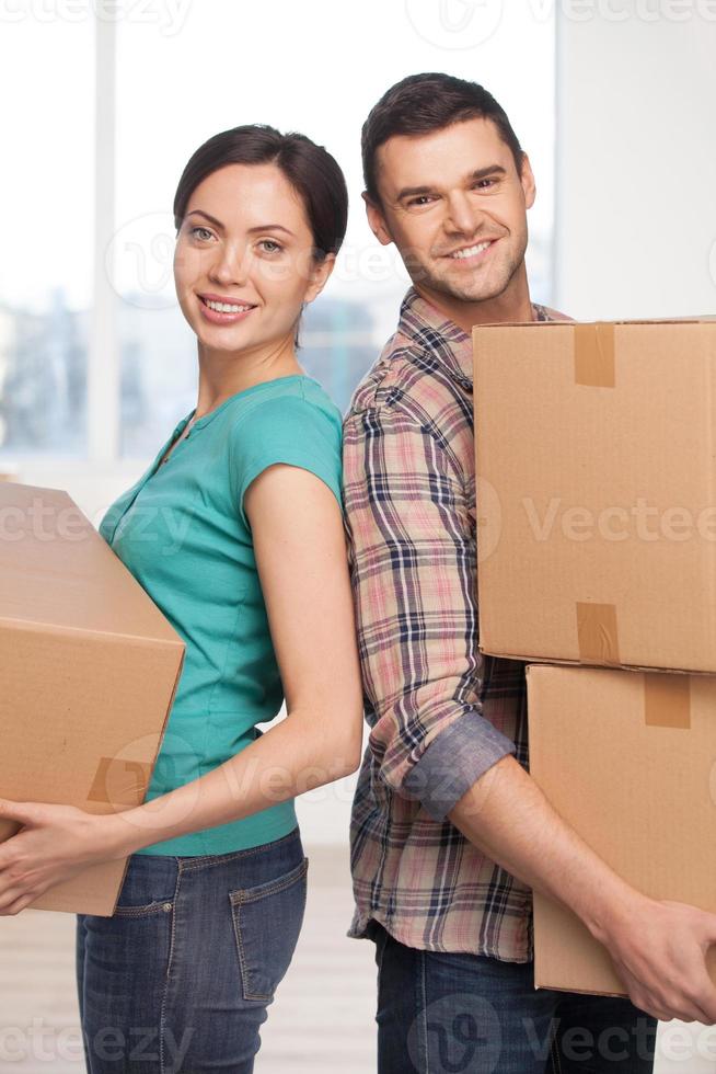 Moving to a new house. Cheerful young couple standing close to each other and smiling at camera while holding cardboard boxes photo