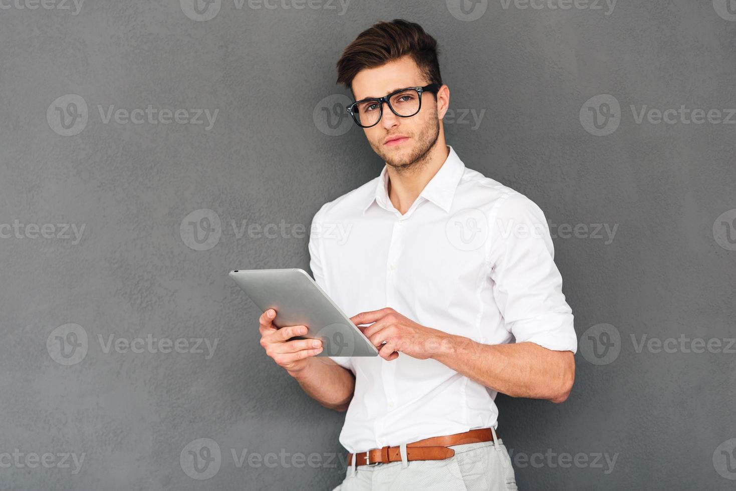 Confident and handsome. Pensive young handsome man using his digital tablet and looking at camera while standing against grey background photo