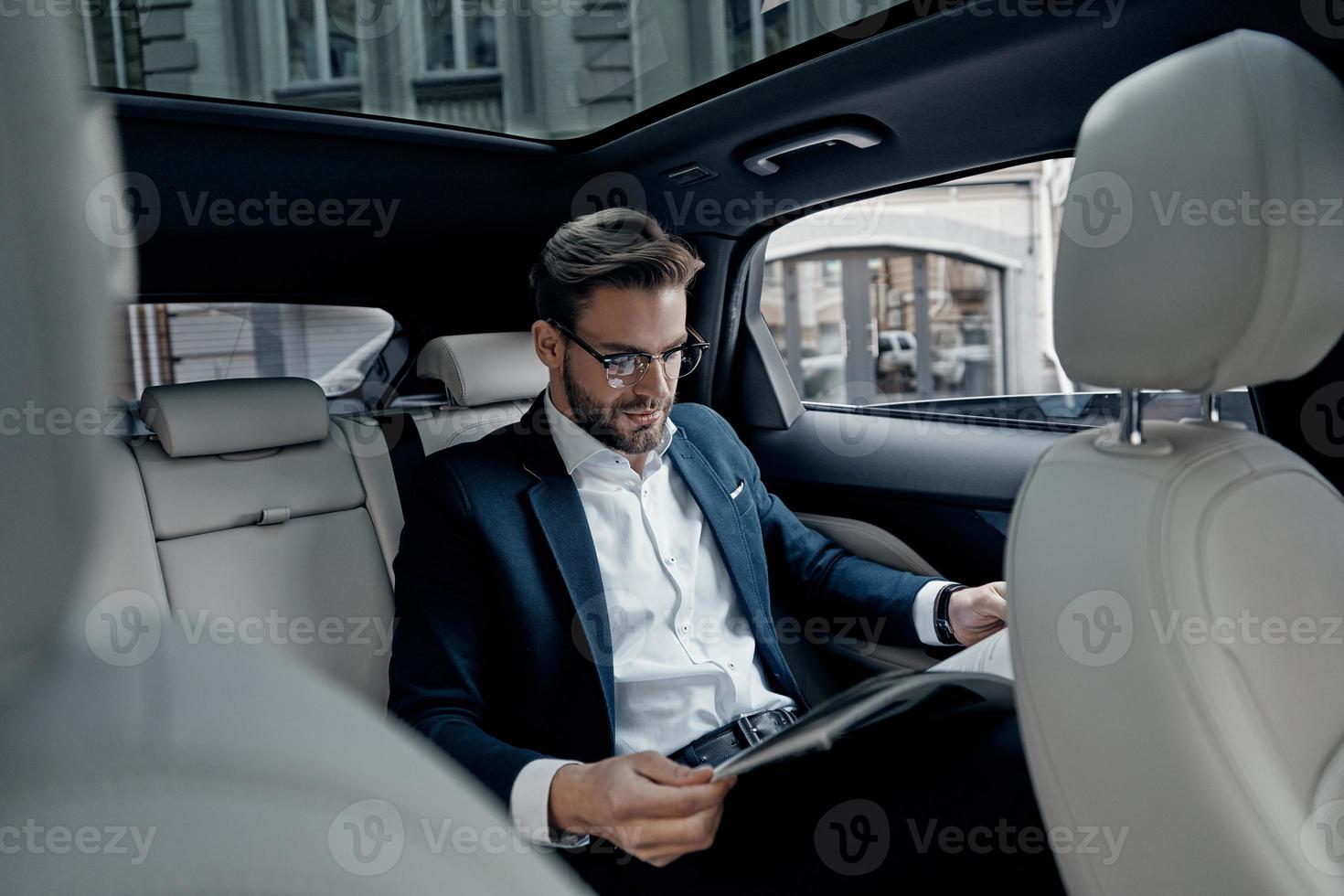 Reading latest news. Handsome young man in full suit reading a newspaper while sitting in the car photo