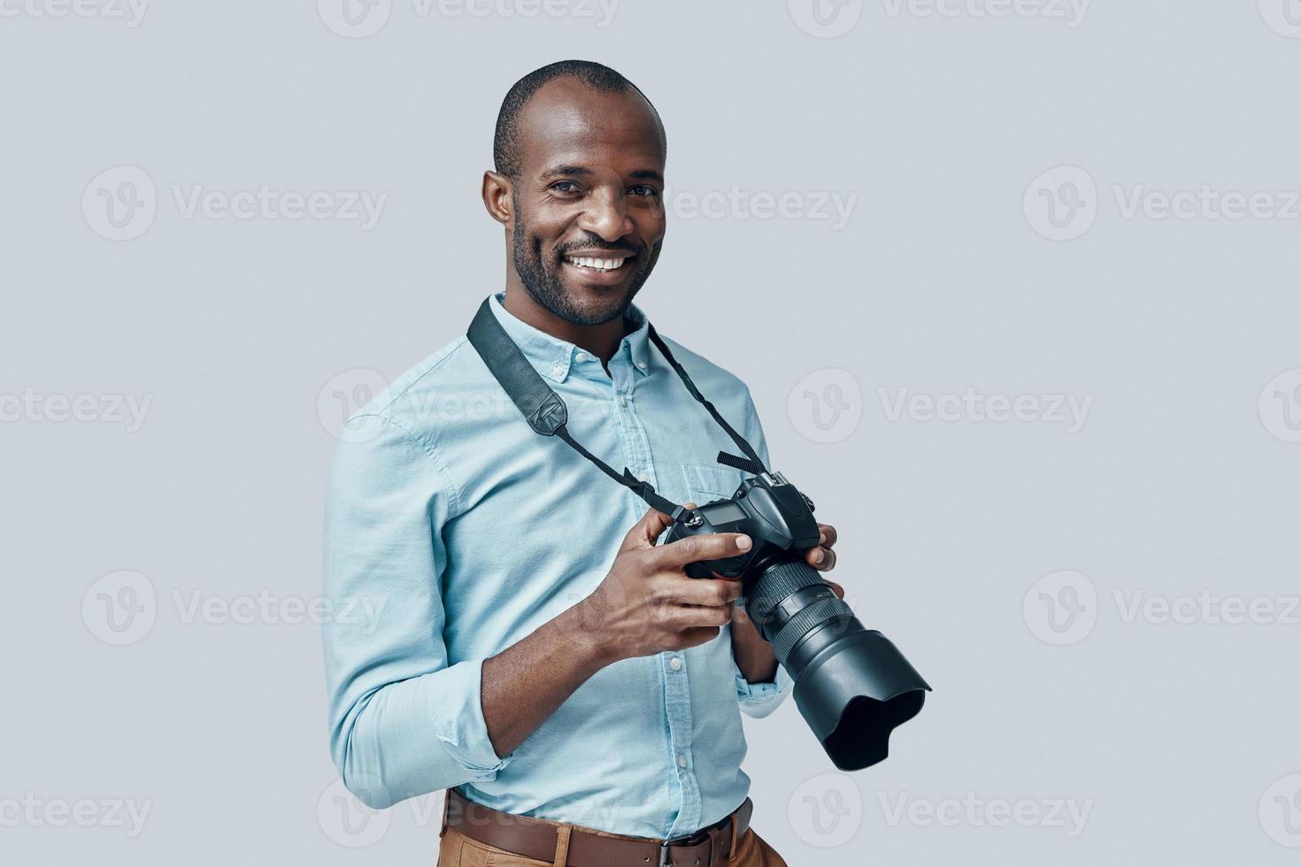 Happy young African man using digital camera and smiling while standing against grey background photo