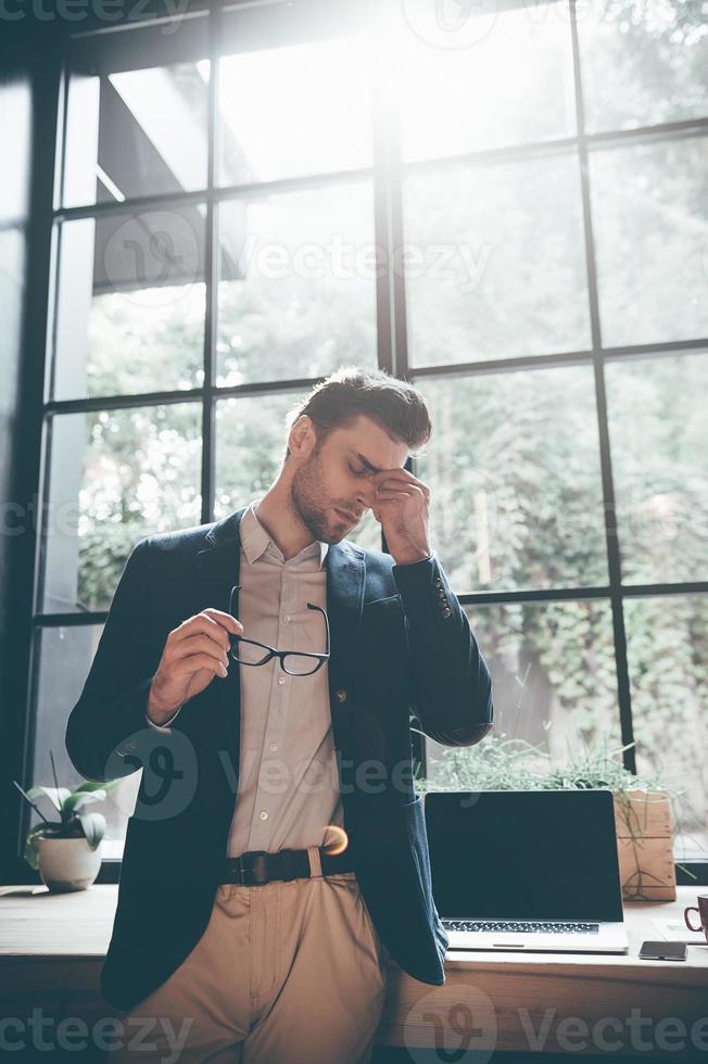 Feeling overworked. Low angle view of frustrated young man carrying eyeglasses and massaging his nose while keeping eyes closed and standing in front of big window in office photo