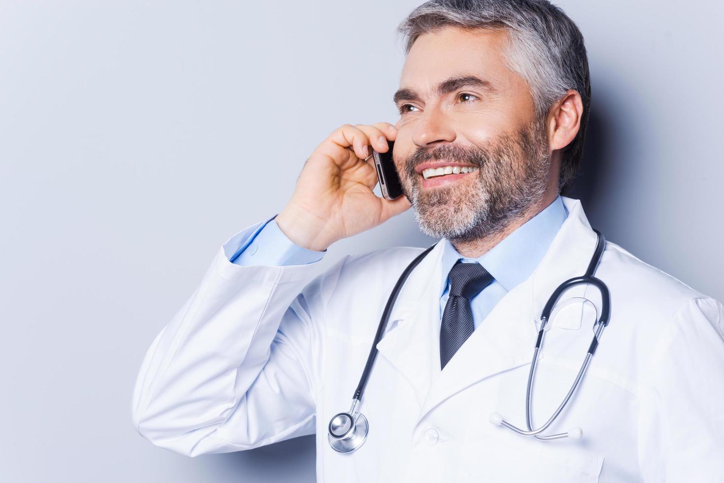 Good news. Confident mature grey hair doctor talking on the mobile phone and smiling while standing against grey background photo