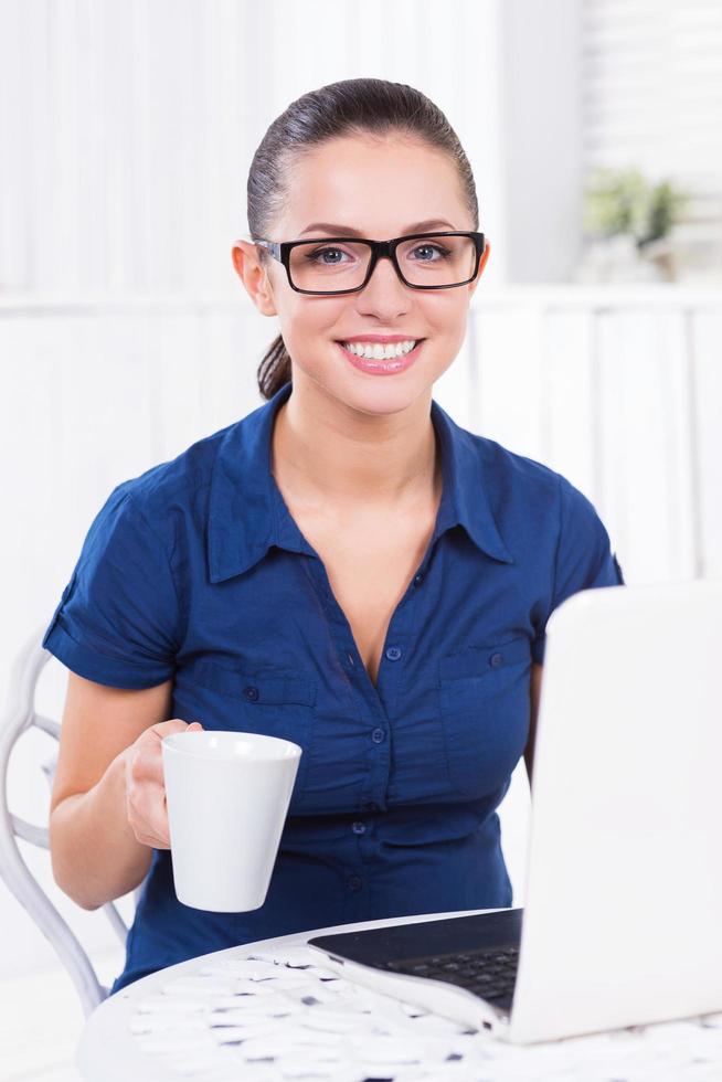 Young and successful. Beautiful young woman working on laptop and holding cup with hot drink photo