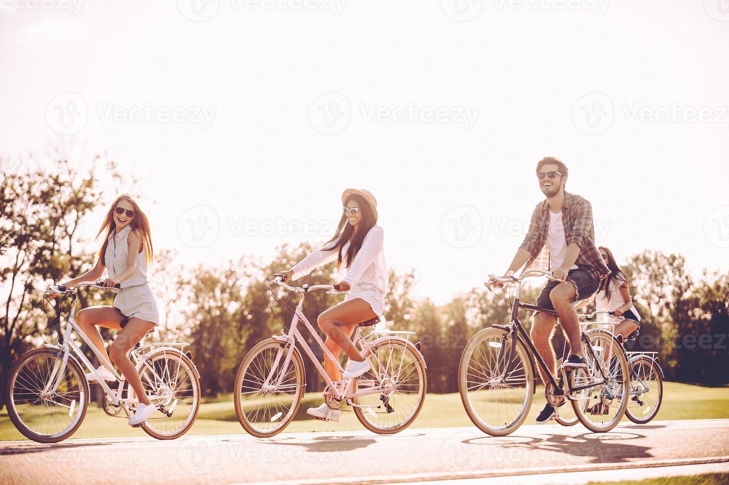 pasar un buen día de verano juntos. grupo de jóvenes montando en bicicleta a lo largo de una carretera y luciendo felices foto