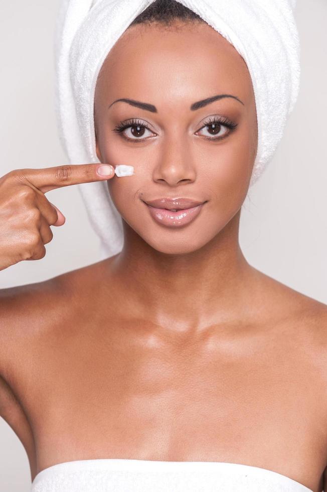 Staying young and beautiful. Close Up image of attractive young Afro-American woman spreading cream on her face and looking at camera while standing against gray background photo