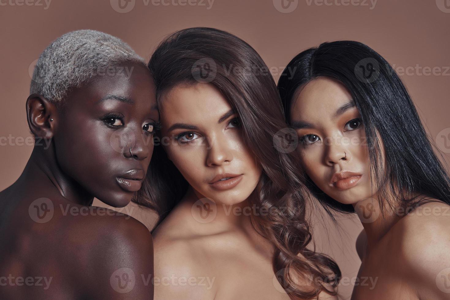 So beautiful Three attractive young women looking at camera while standing against brown background photo