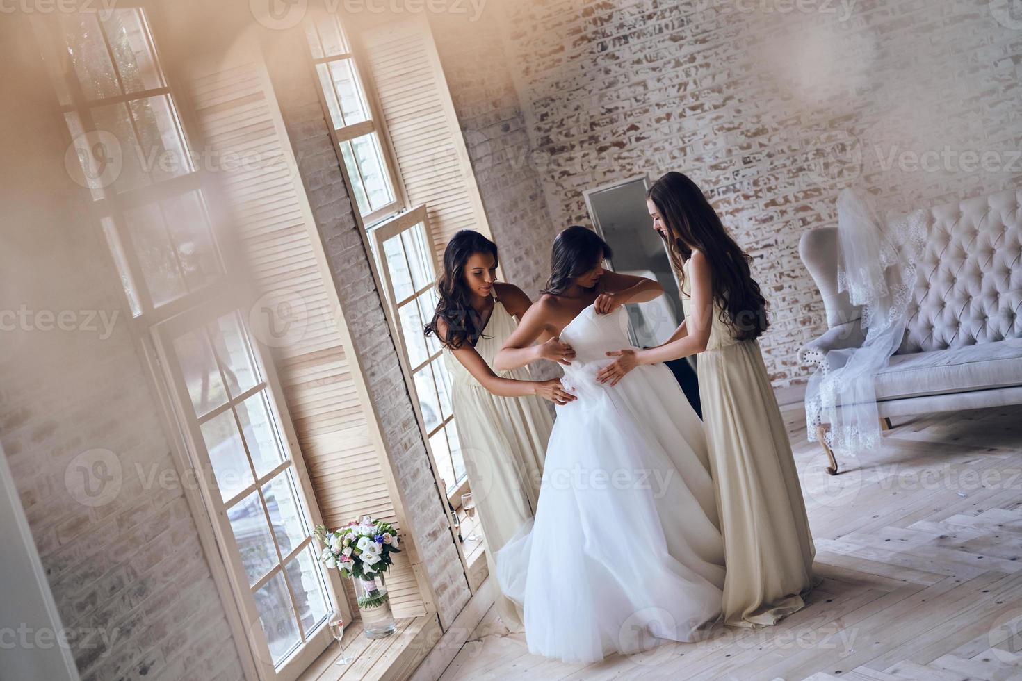 Important preparations. Full length top view of two attractive young women putting on a wedding dress on a bride while standing neat the window together photo