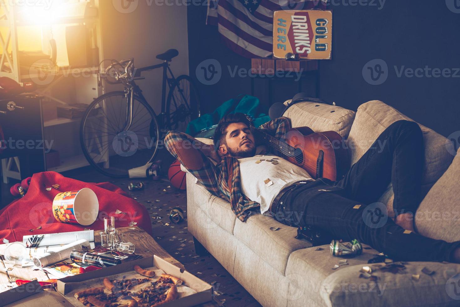 Little rest can help with hangover. Young handsome man lying down on sofa and napping in messy room after party photo