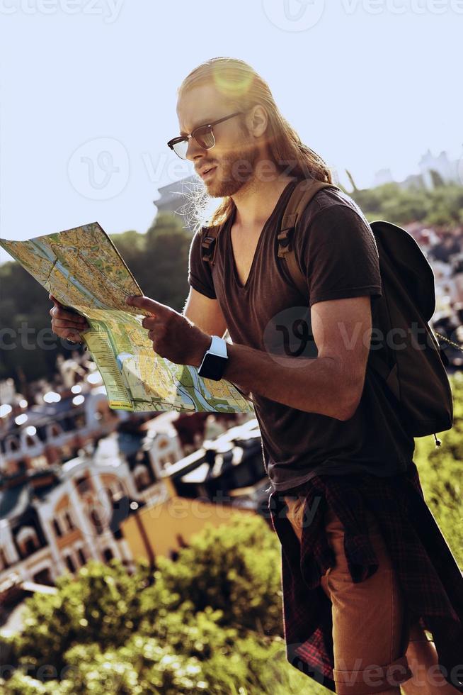 Where to go next Handsome young man in casual clothing looking at map while standing on the hill outdoors photo