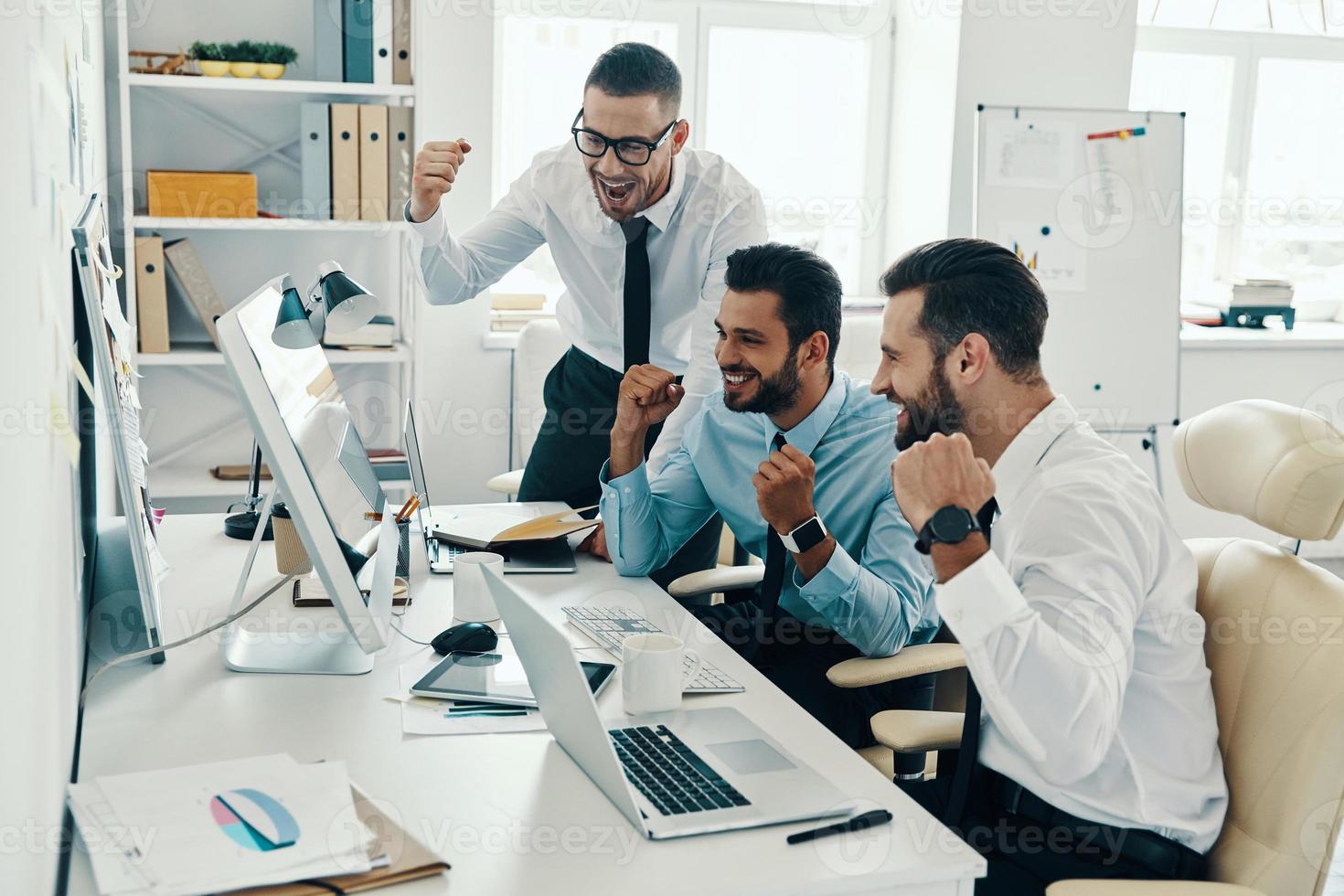 Finally success. Group of young modern men in formalwear smiling and gesturing while working in the office photo