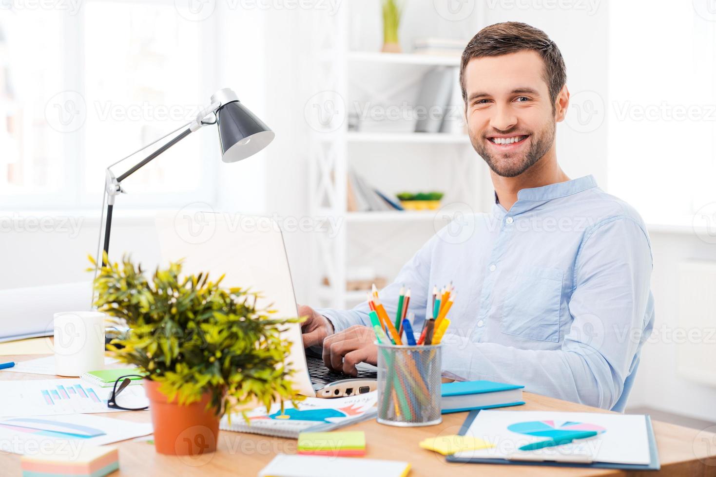 trabajando con placer. un joven guapo con camisa que trabaja en una laptop y sonríe a la cámara mientras se sienta en su lugar de trabajo foto