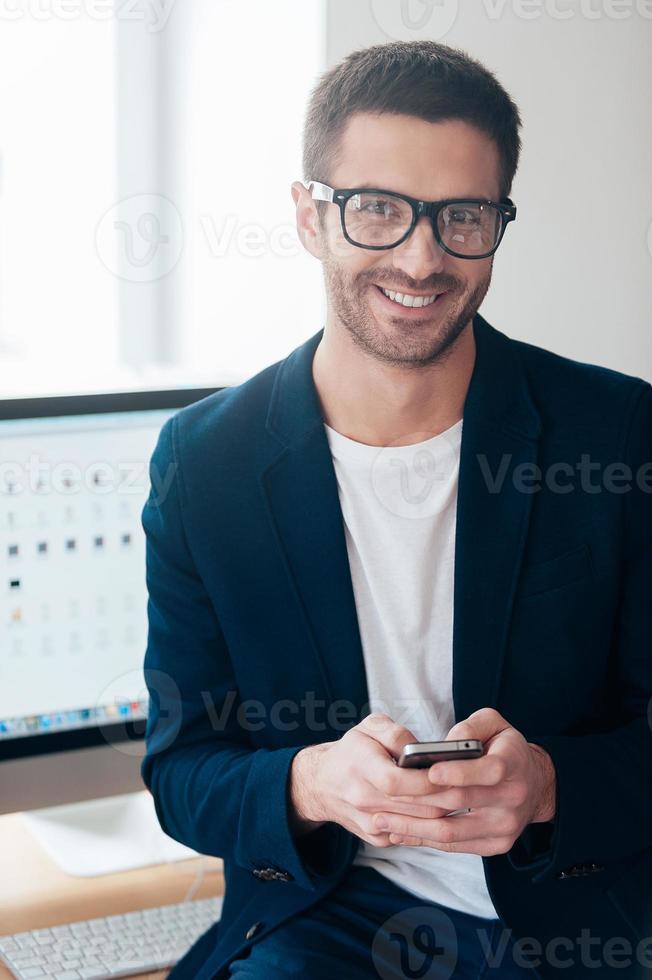 Smart and confident. Confident young man holding smart phone and smiling while leaning at the office desk photo