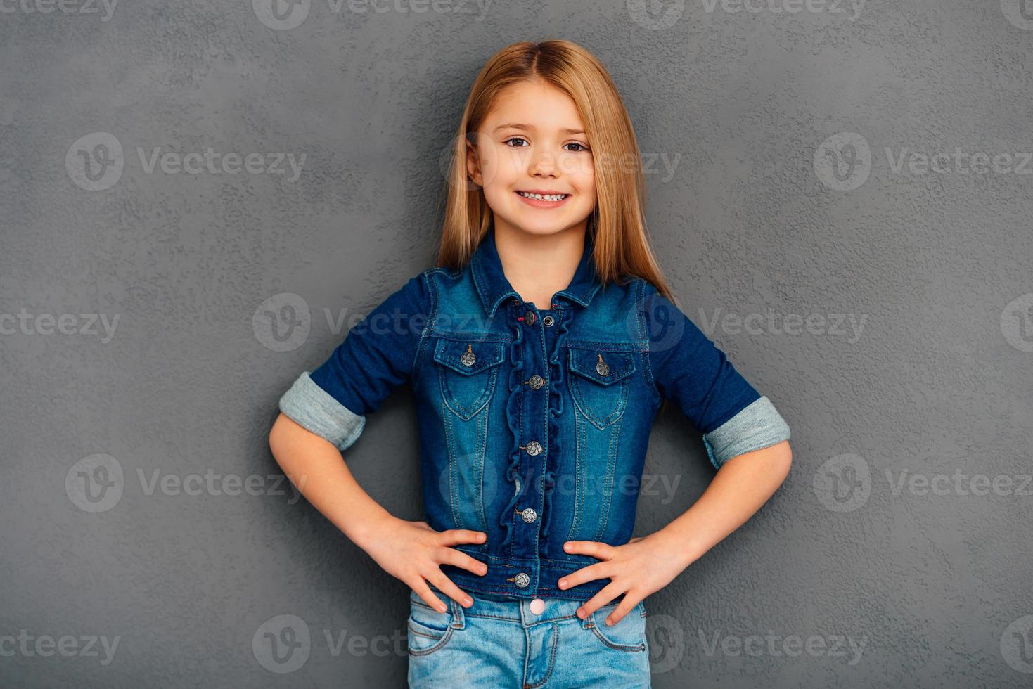 Confident and smiley girl. Cheerful little girl holding hands on hips and looking at camera with smile while standing against grey background photo