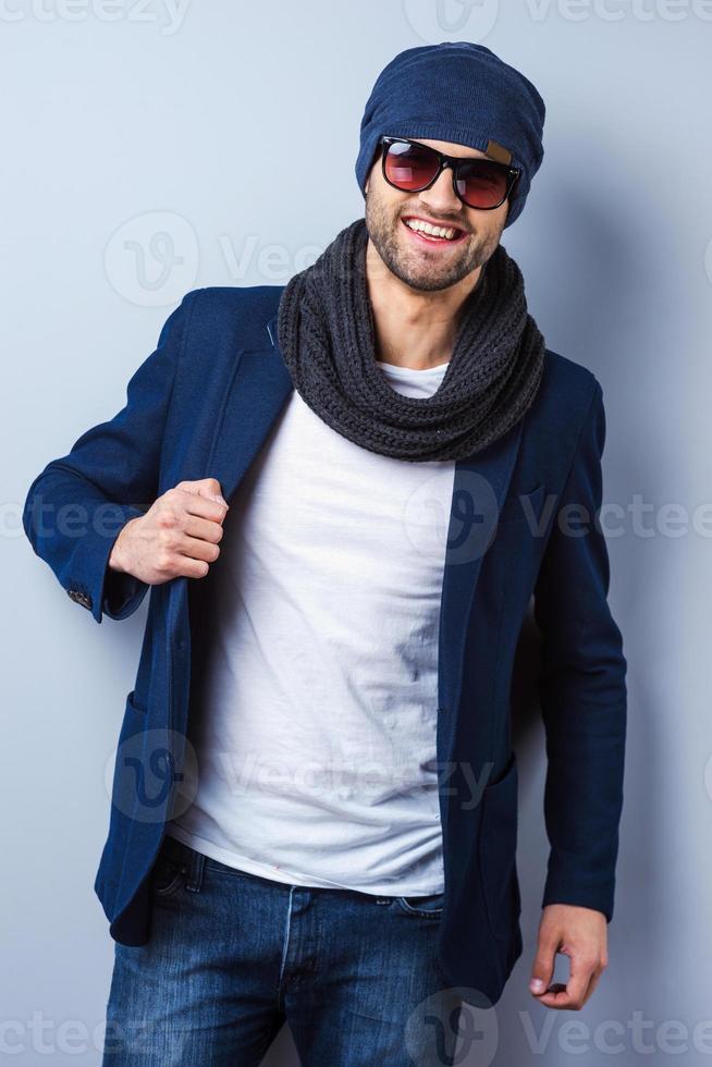 Confident in his perfect style. Handsome young stylish man in sunglasses and hat adjusting his jacket and looking at camera while standing against grey background photo