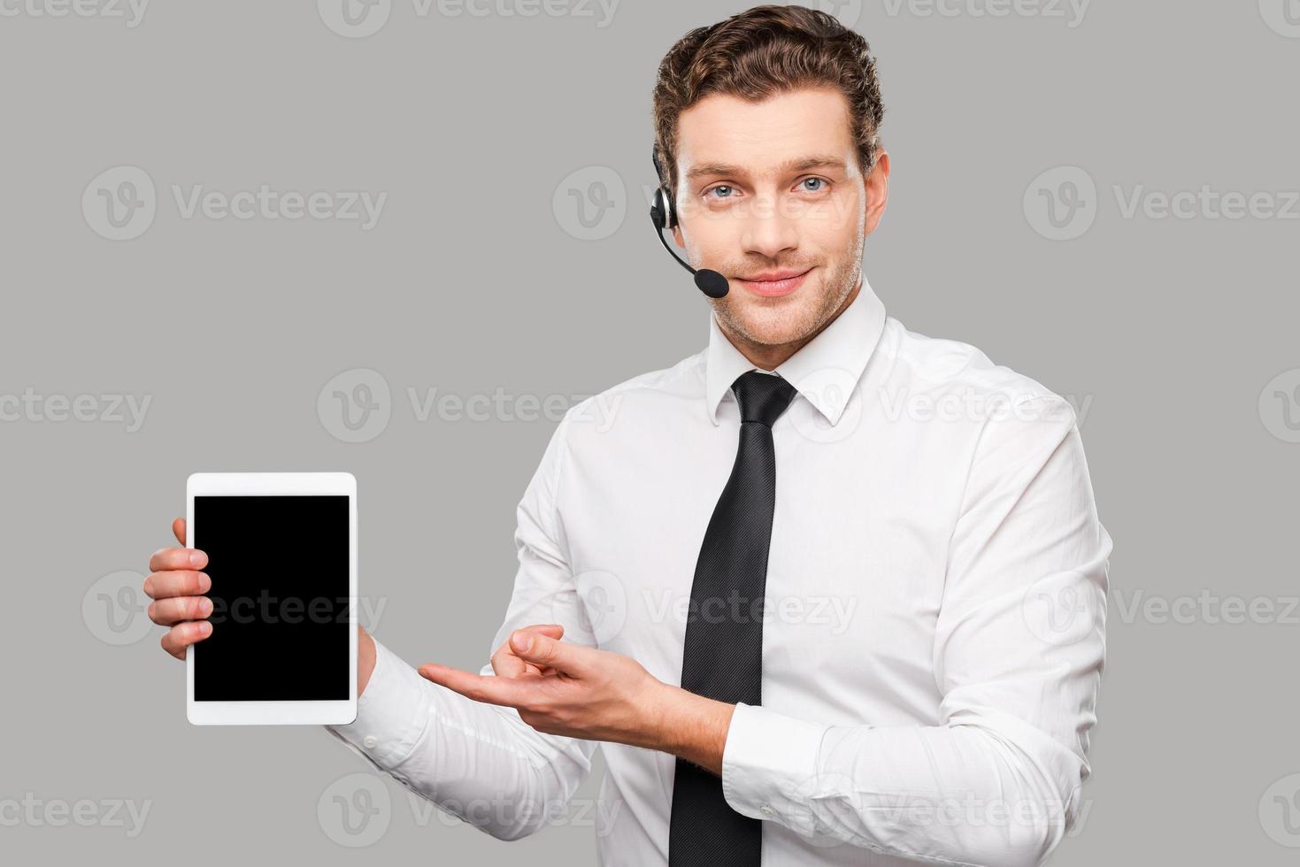Copy space on his laptop. Handsome young man in formalwear and headset pointing  digital tablet while standing against grey background photo