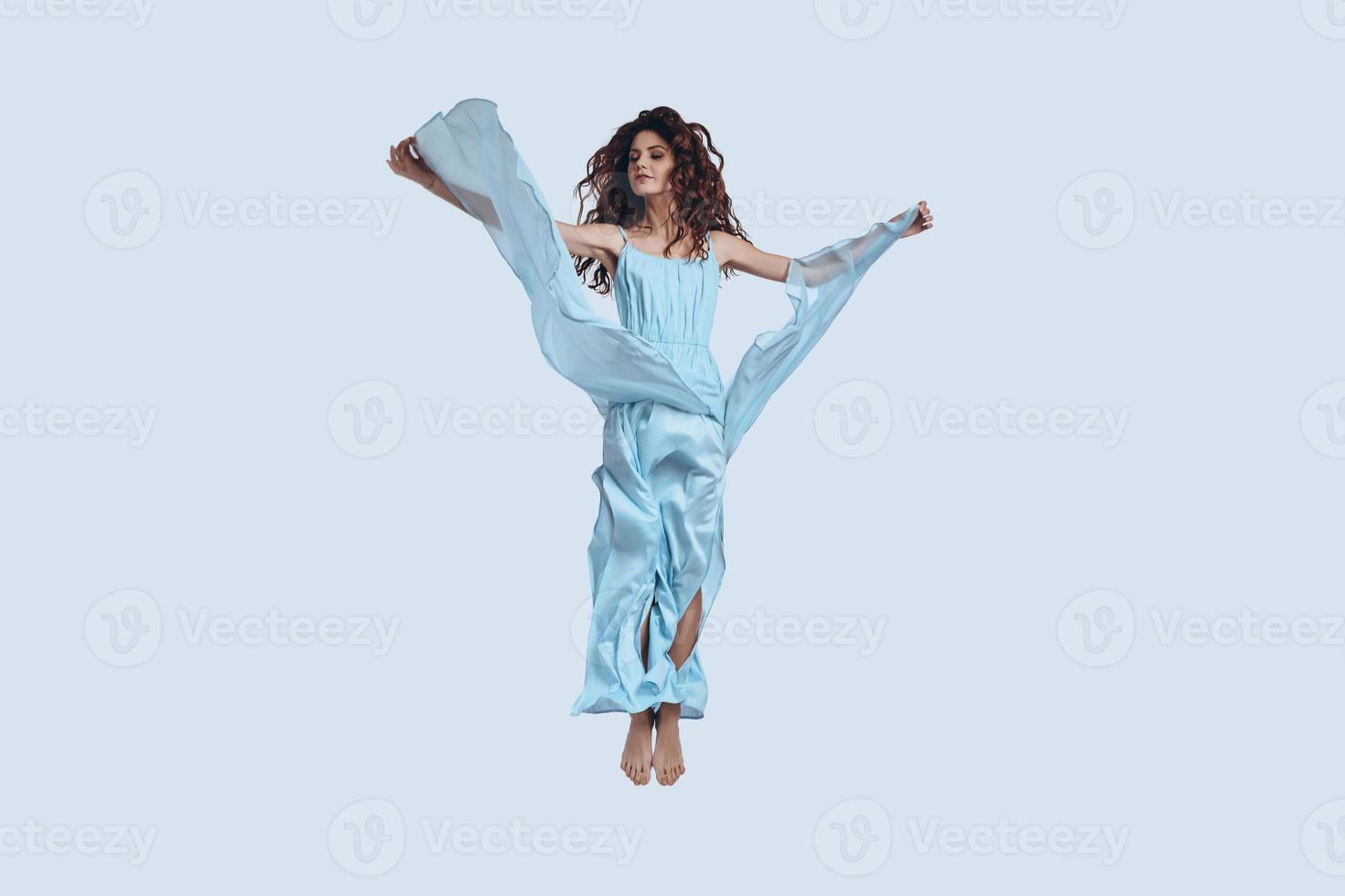 Flying high.... Full length studio shot of attractive young woman in elegant dress gesturing while hovering against grey background photo